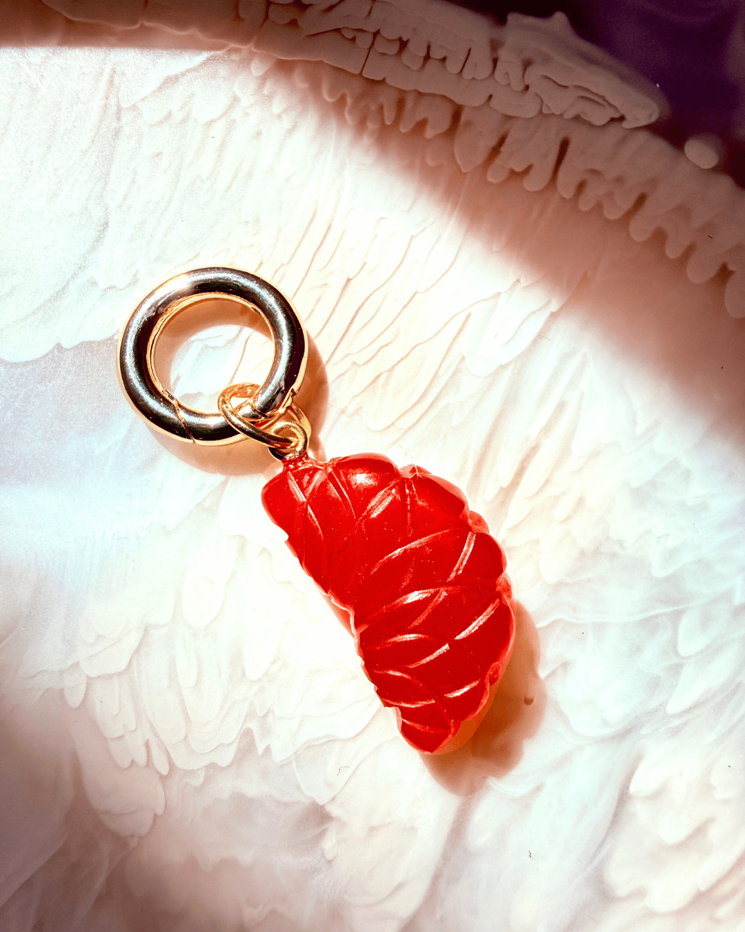 An orange carnelian jube charm with a gold-tone ring and connector, resting on a textured white surface under dramatic warm lighting.