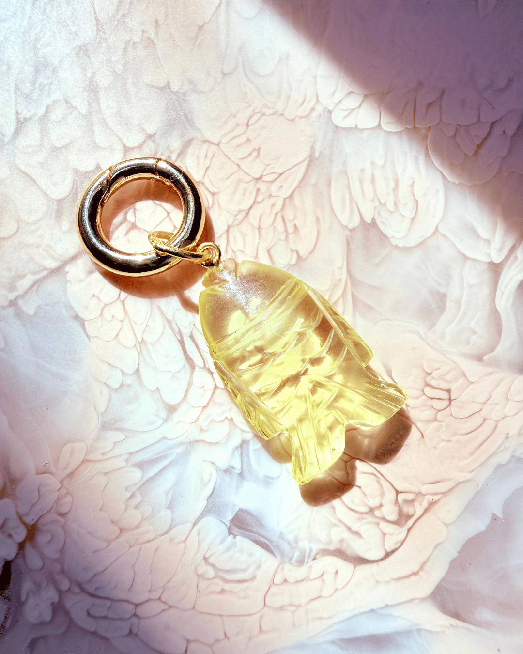 A gold-toned metal key ring with a translucent pale lemon quartz fish charm shaped like a small figure, photographed on a textured white surface with strong sunlight and shadows.