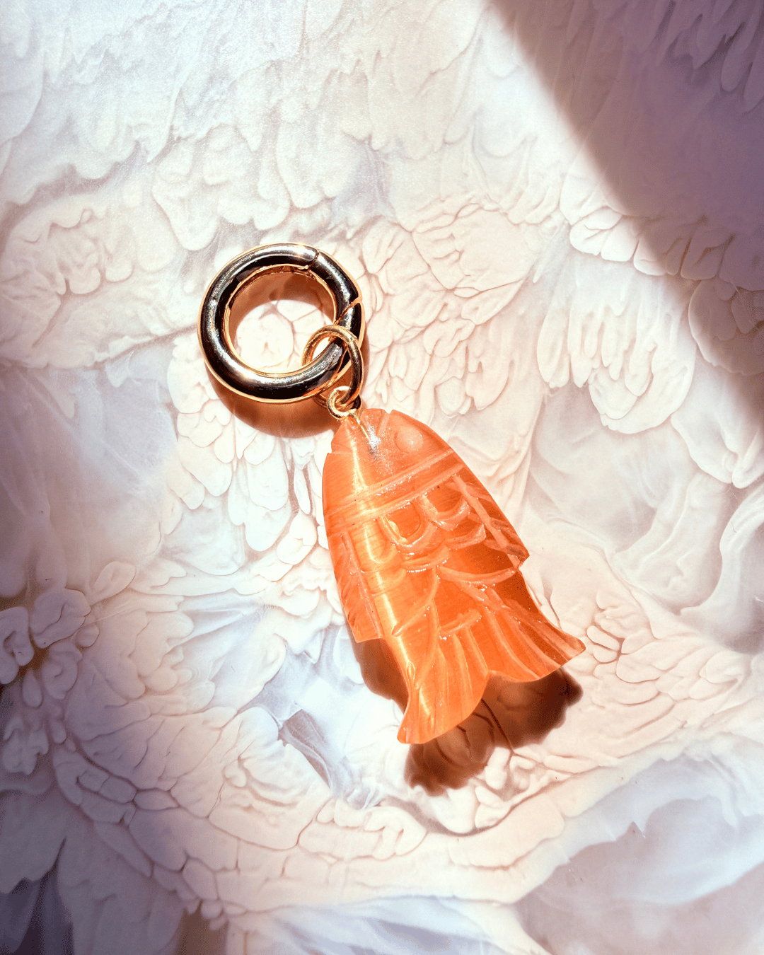An orange translucent fish-shaped pendant charm with carved details, attached to a gold-tone split ring, resting on a textured white fabric background.
