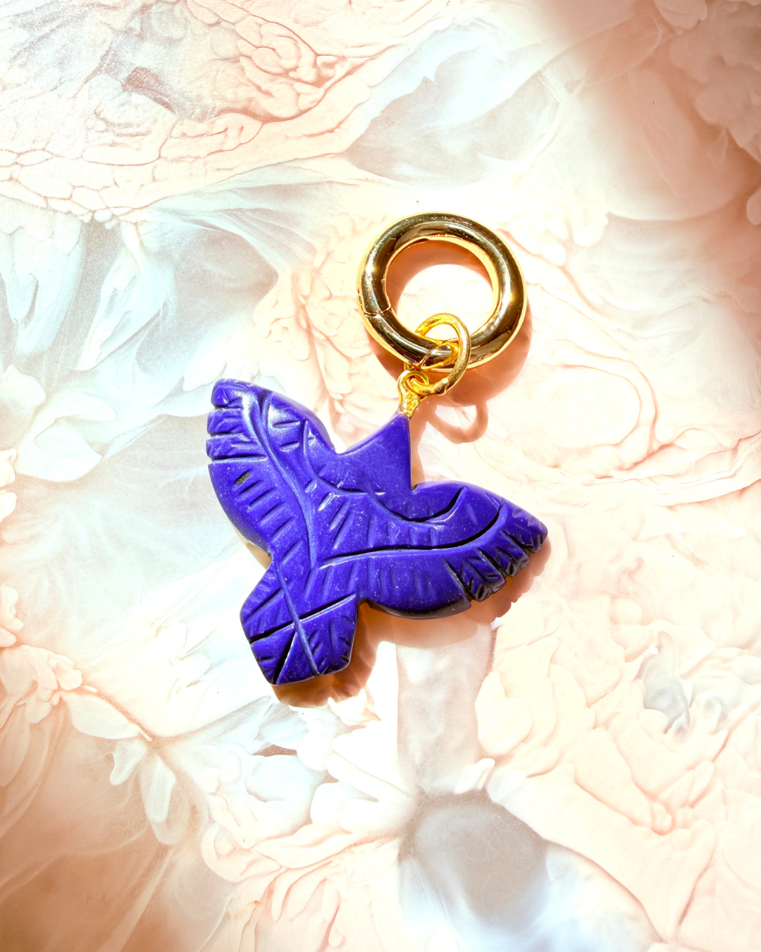 A bluecarved bird charm with a gold metal ring clasp, resting on a textured, light pink and white marbled surface.