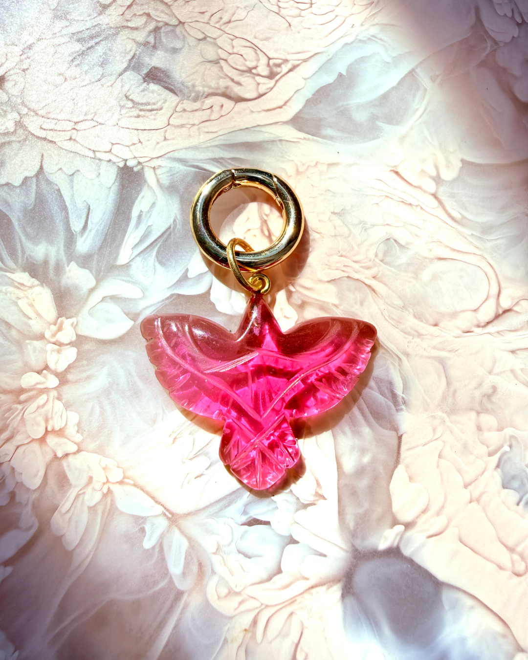 A pink bird-shaped charm with a gold-tone ring clasp, resting on a textured, light pink and white floral surface.