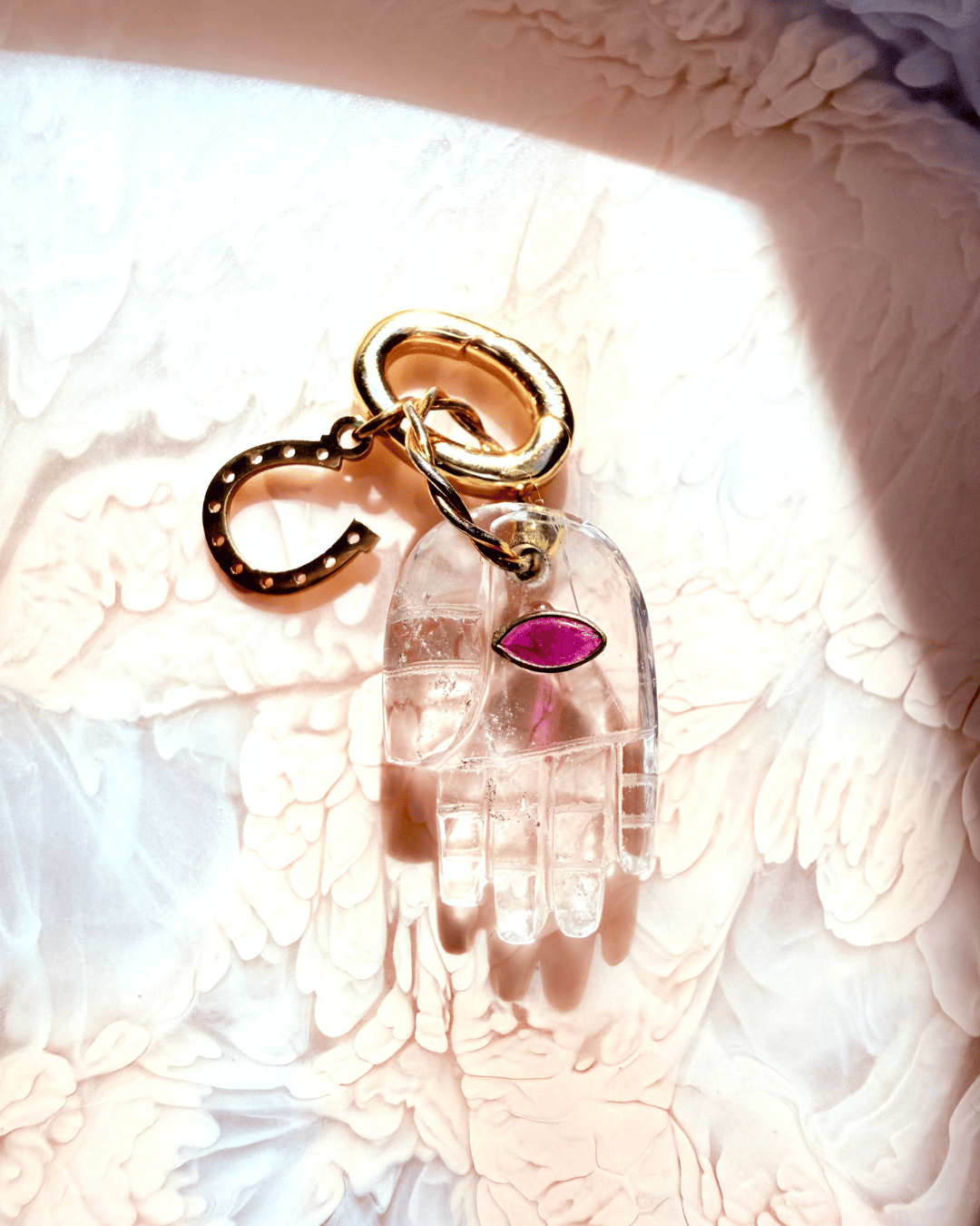 A clearrock crystal Hamsa hand keychain charm with a magenta-pink eye and gold-tone metal hardware, resting on a textured white surface.