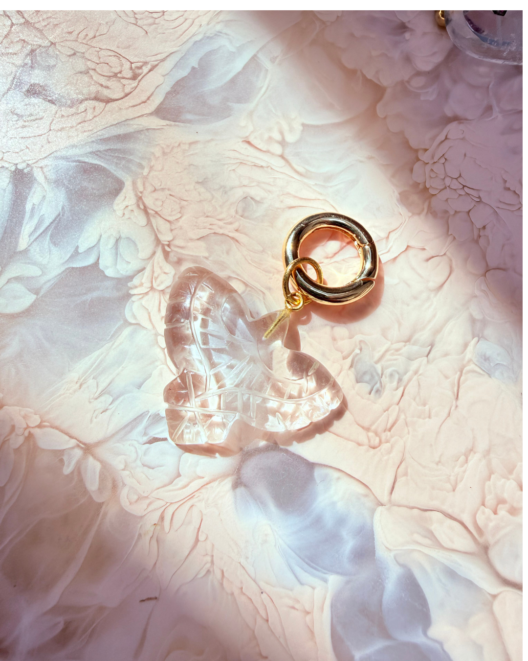A clear bird-shaped charm with a gold ring clasp, resting on a textured, marbled pink and white surface.
